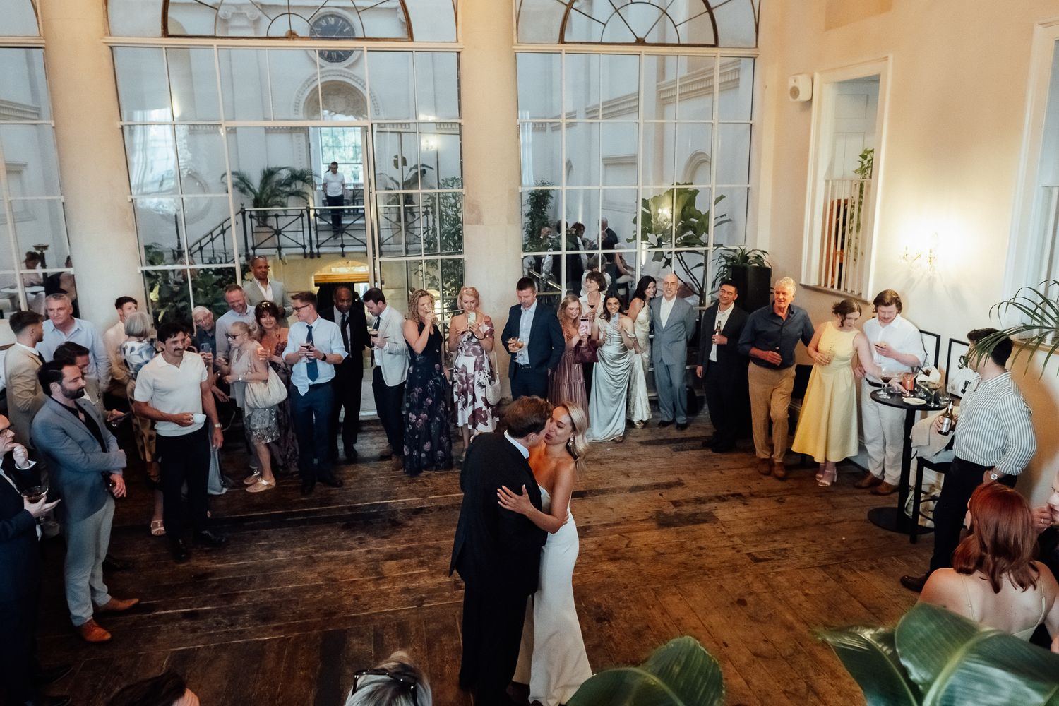 Newlyweds share a first dance on a wooden floor as wedding guests watch in a bright reception hall with tall arched windows.
