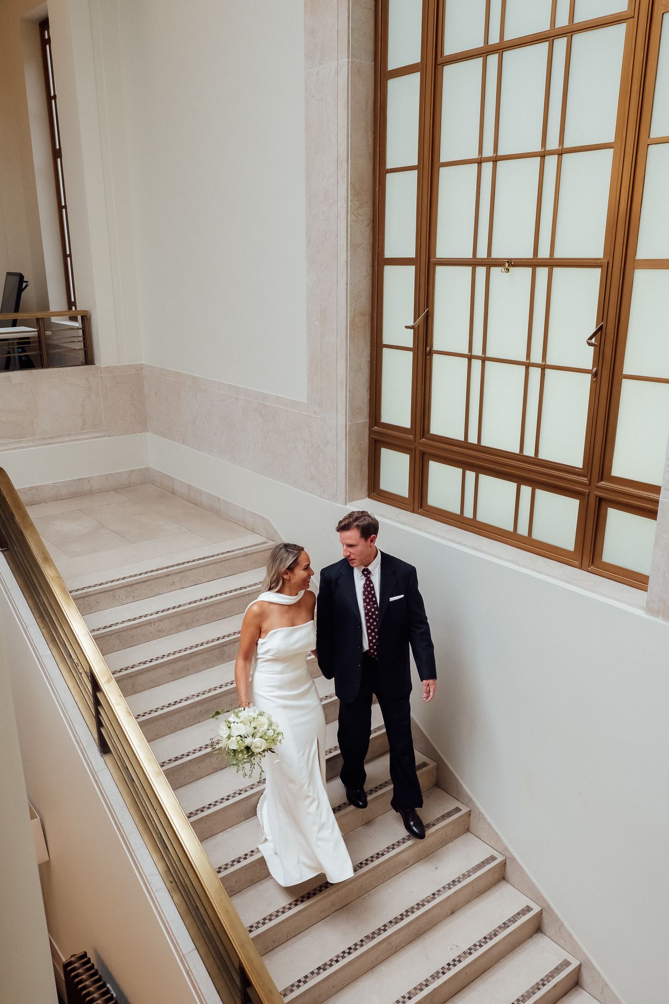 Bride in a white gown holding a bouquet walks down a marble staircase beside a groom in a navy suit, near a large window.