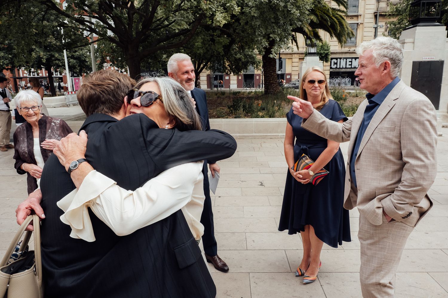 Two women share a warm hug outdoors while an older man in a beige suit gestures and smiles, with guests gathered nearby at a wedding.