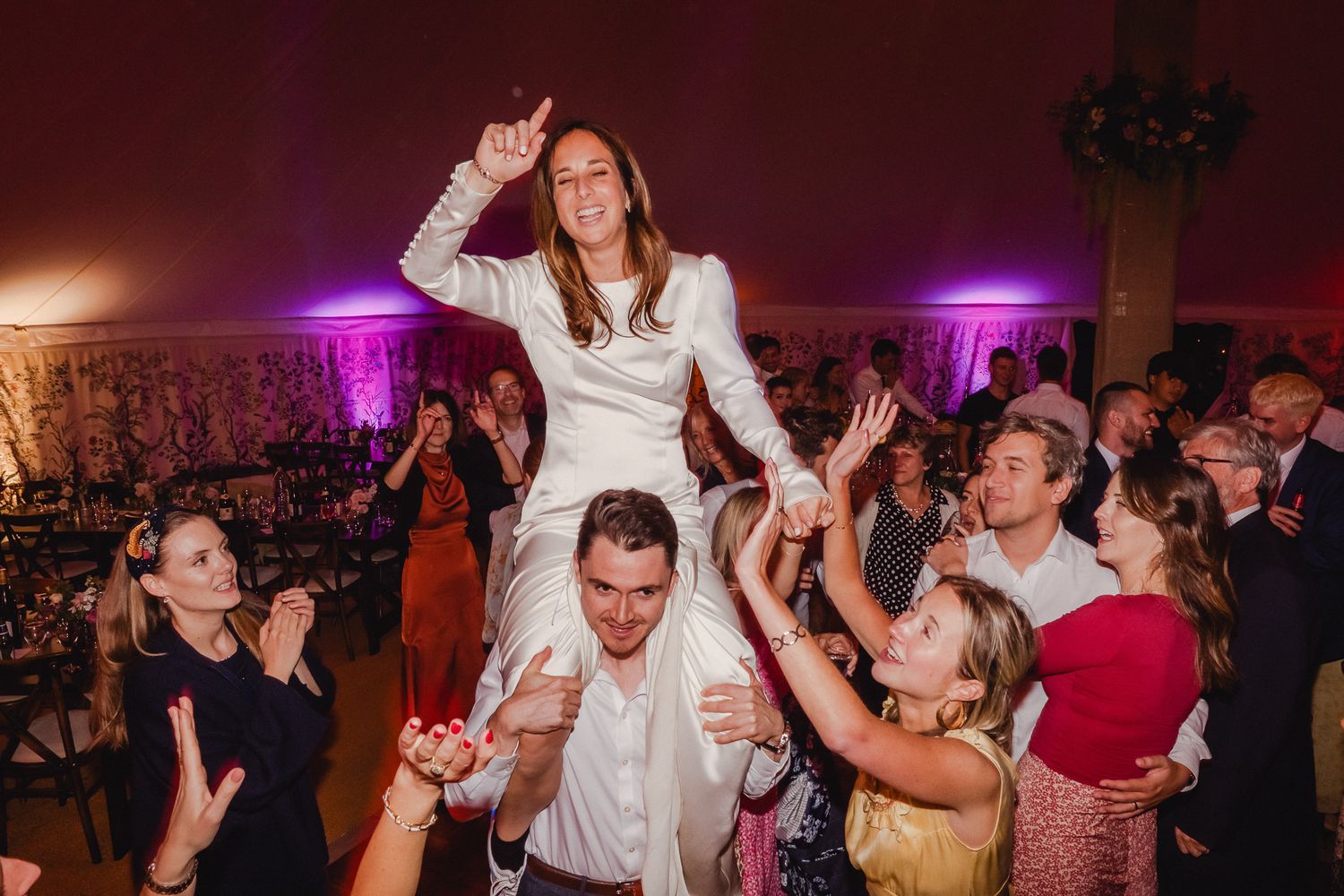 Wedding guests dance and celebrate on a crowded reception dance floor with purple lighting in the background.