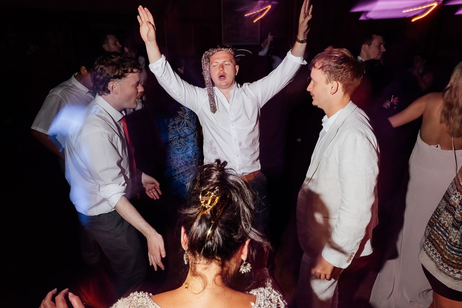Guests dancing energetically at a wedding reception with hands raised in celebration under colorful lights.