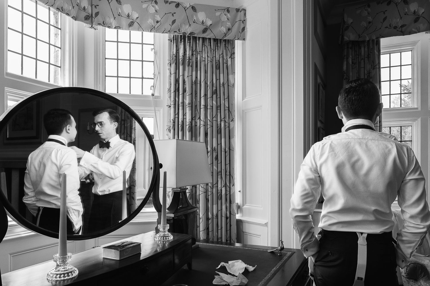 Black and white photo of a groom adjusting his bow tie while reflected in a round mirror in an elegant room.