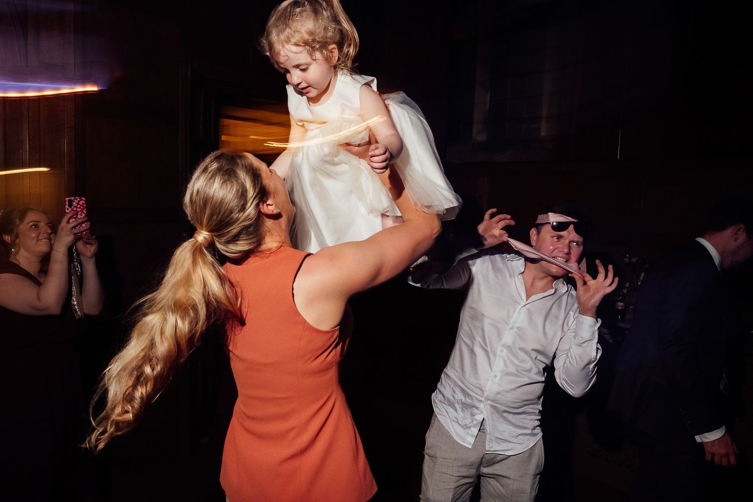 Woman in orange dress lifting young girl in white at a lively wedding reception dance floor.