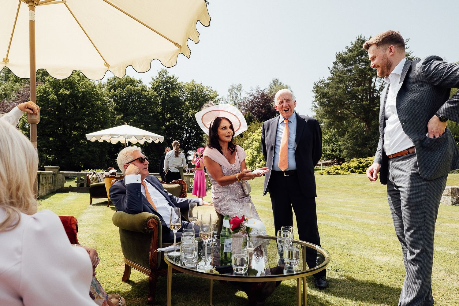 Elegant garden party guests socializing outdoors on sunny day, woman in white hat, men in suits near drinks table.