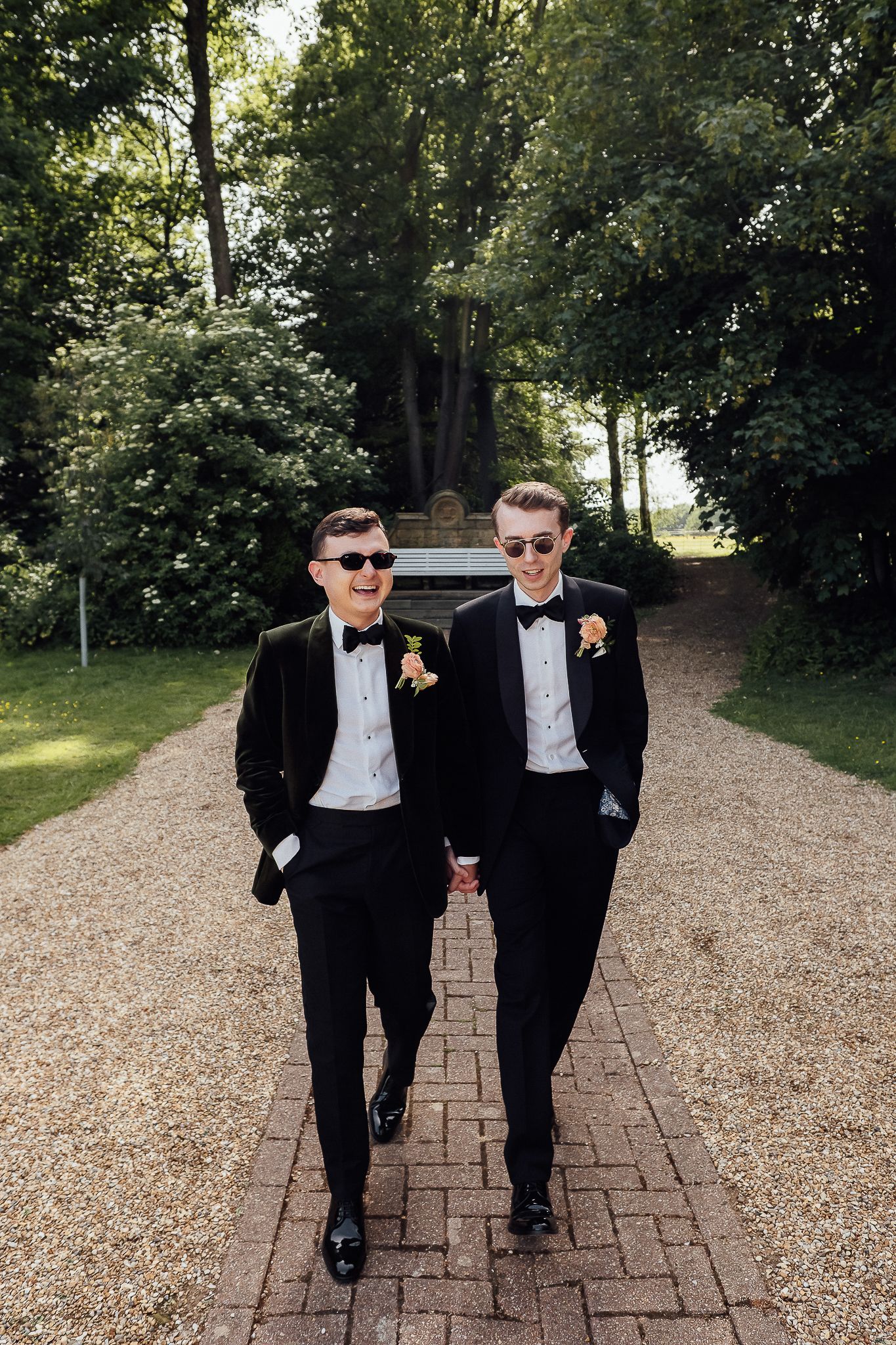 Two grooms in black tuxedos and sunglasses hold hands while walking down a garden path on their wedding day.