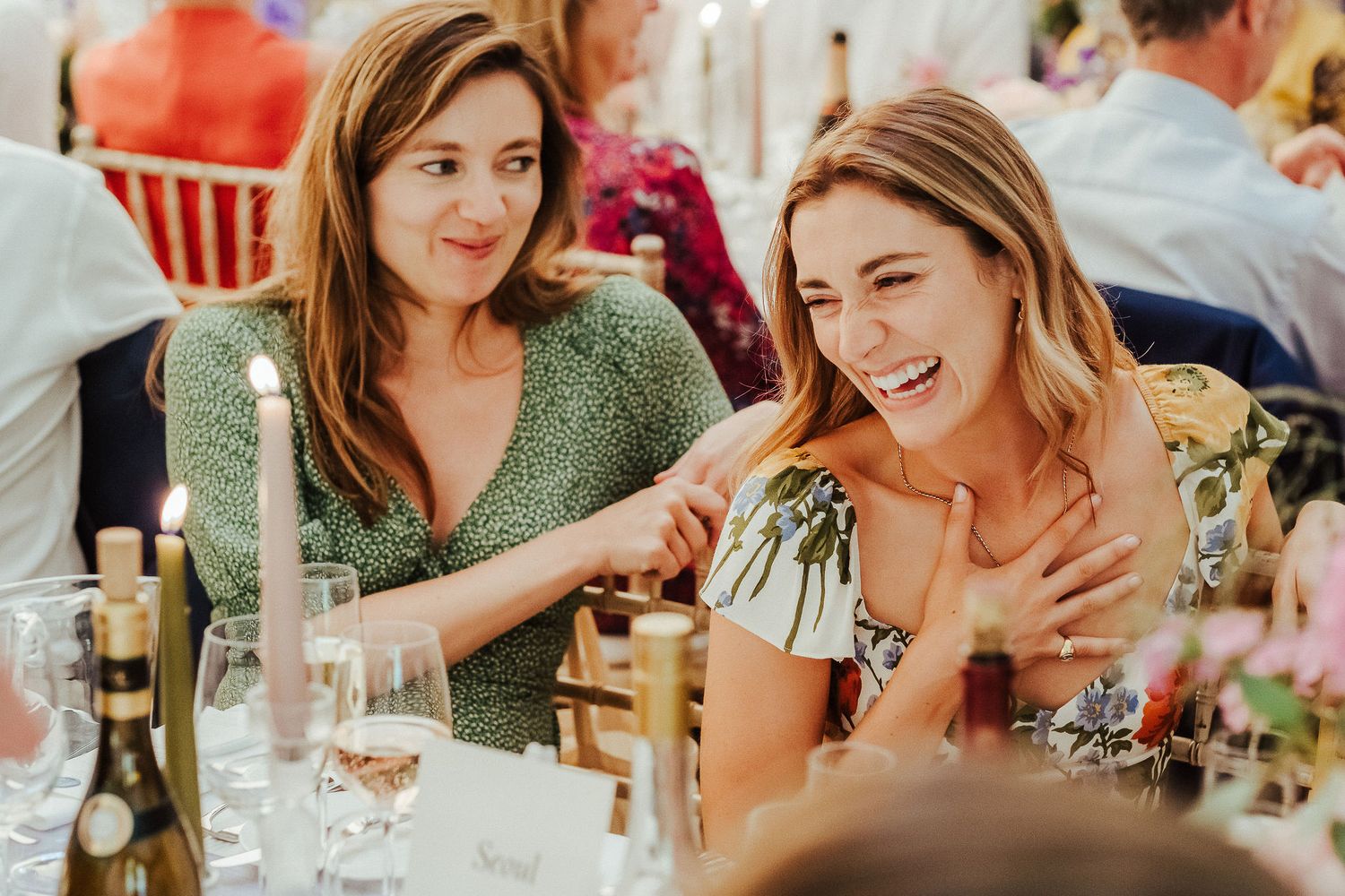 Two wedding guests in colorful dresses share lighthearted moment at reception dinner table.