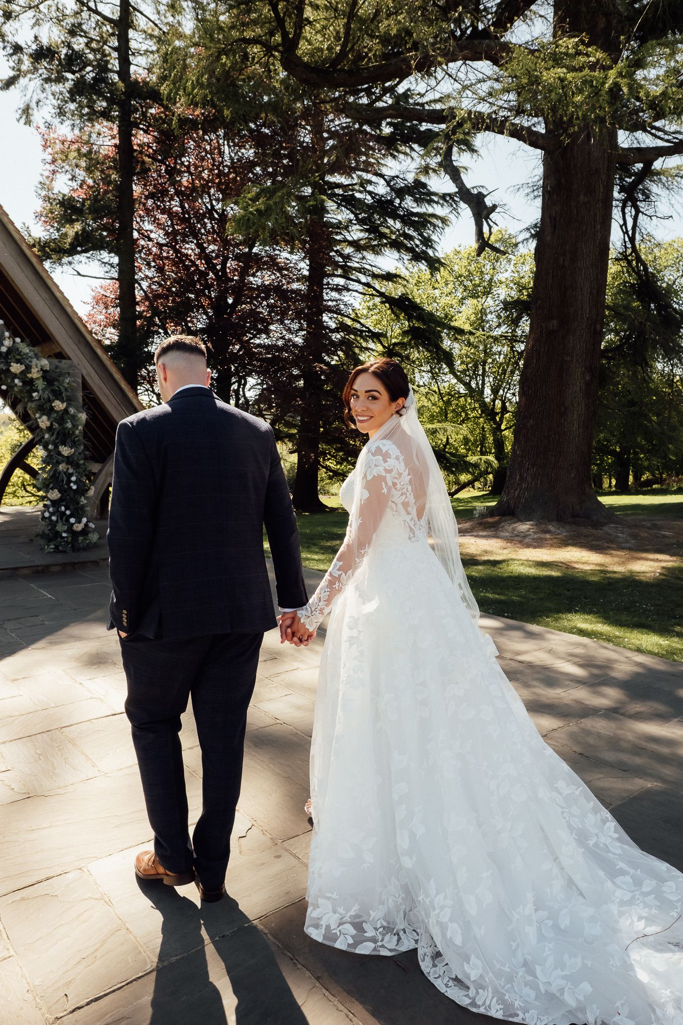 Bride in lace gown and groom in dark suit walk hand-in-hand through a sunlit garden on their wedding day.
