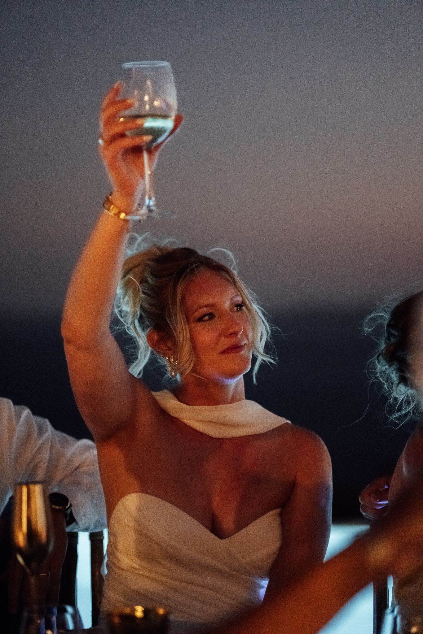 Bride in white strapless gown raises a wine glass in a toast at an elegant outdoor evening wedding reception.