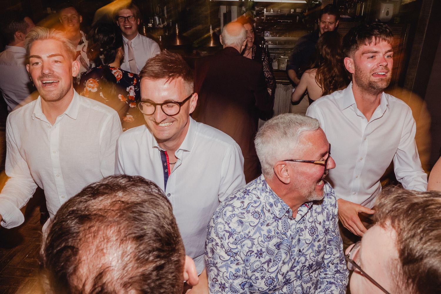 A lively wedding reception shows guests in white shirts and floral prints gathered around tables in a dimly lit venue.