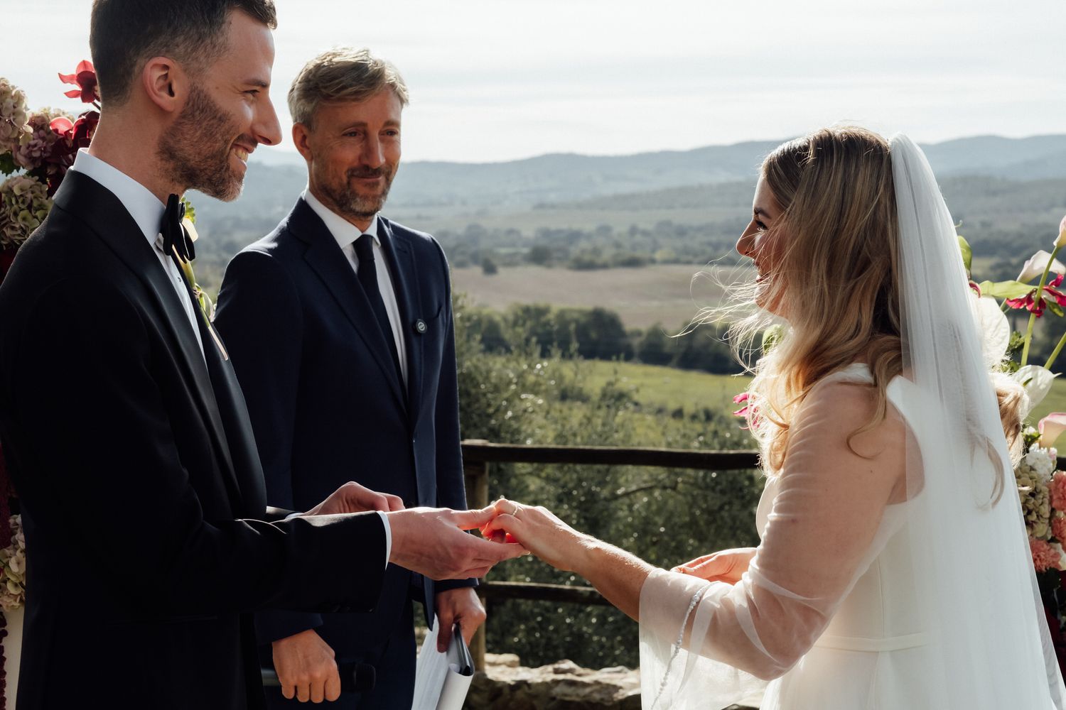 Bride in a white veil and groom in a dark suit exchange rings during an outdoor ceremony with an officiant and scenic hills behind them.