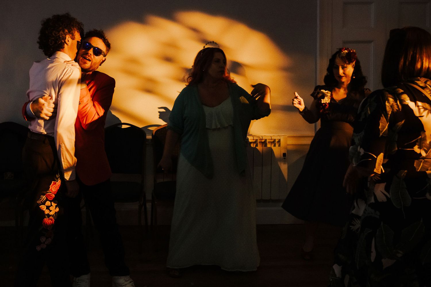 Wedding guests dance and laugh in a dimly lit room as warm golden sunlight casts dramatic patterns across the wall behind them.