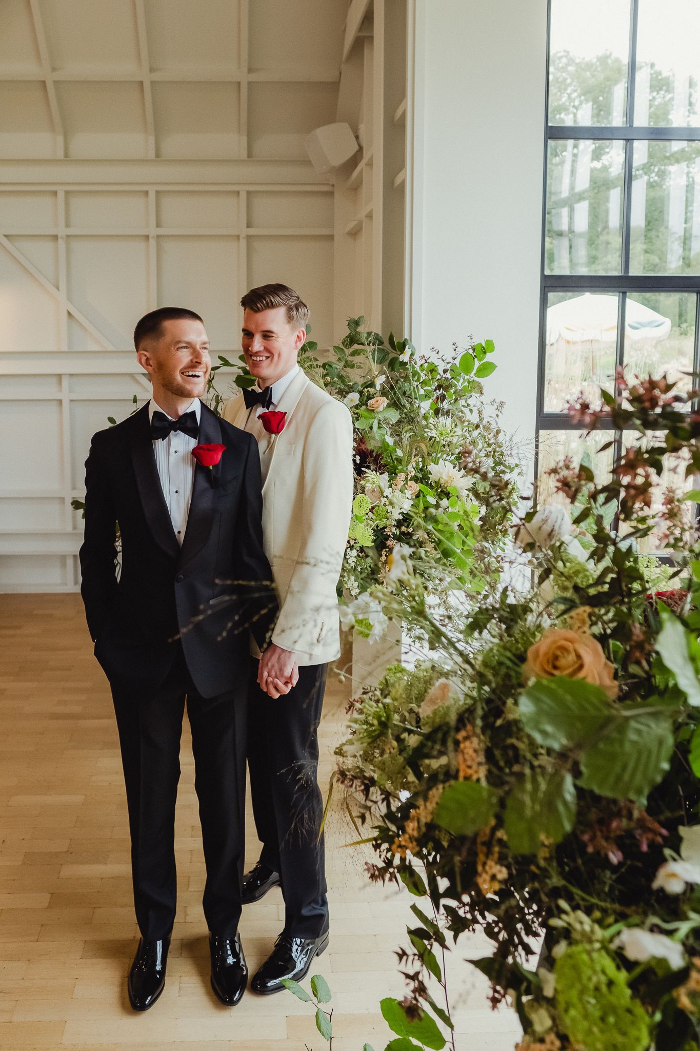 A couple in formal attire shares a romantic moment beside elegant floral arrangements in a bright indoor venue.