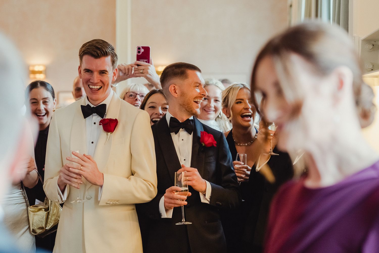 A group of wedding guests laugh and smile while holding drinks during a reception celebration.