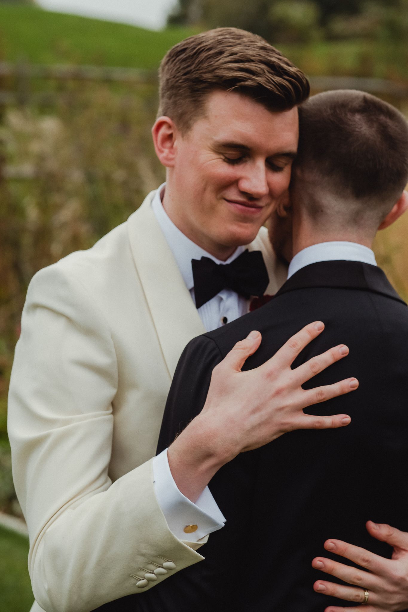 Two people in formal attire embrace during a wedding ceremony, one wearing a white tuxedo and black bowtie.