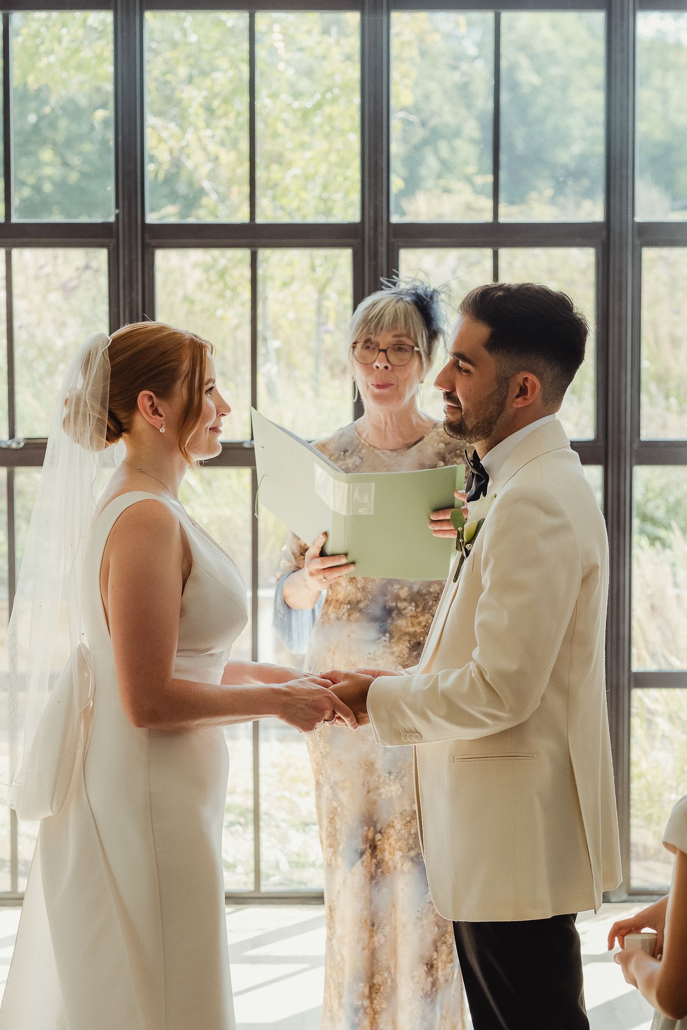 Wedding party in white attire gathered by large windows during indoor celebration.