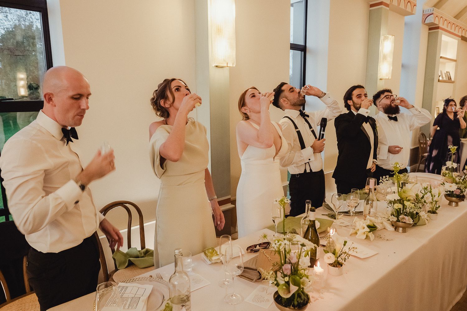 Wedding party members share laughter and conversation while seated at an elegant white reception table with floral centerpieces.