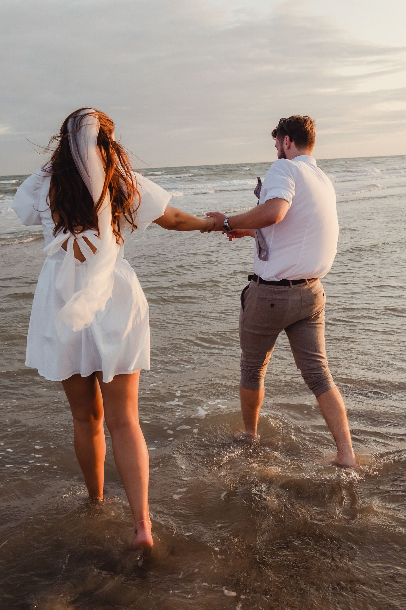 A couple holding hands while walking and splashing in the ocean waves at sunset wearing white and brown summer outfits.