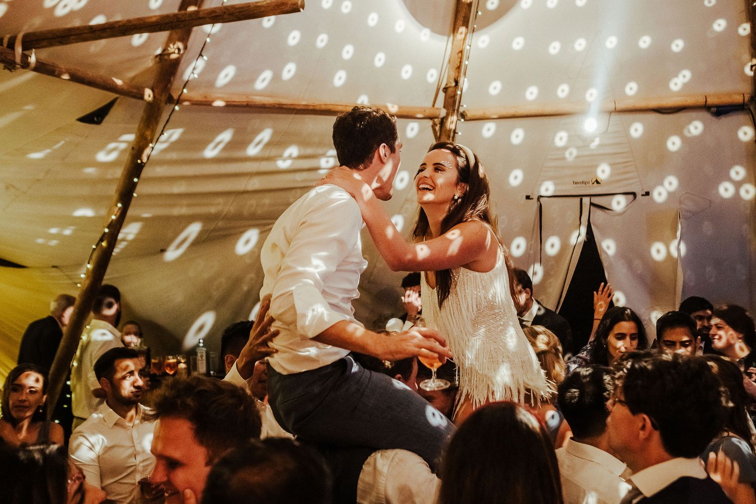 Newlyweds celebrate on shoulders during reception under string-lit tent with excited guests below.