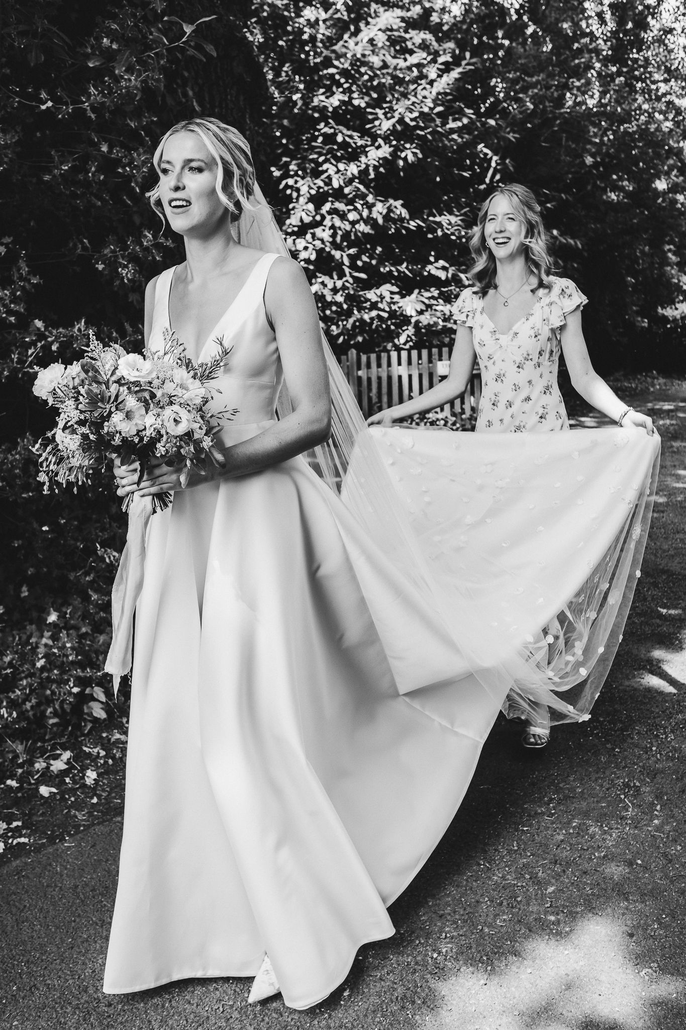 Black and white photo of a bride in white gown walking with bridesmaid in floral dress outdoors.
