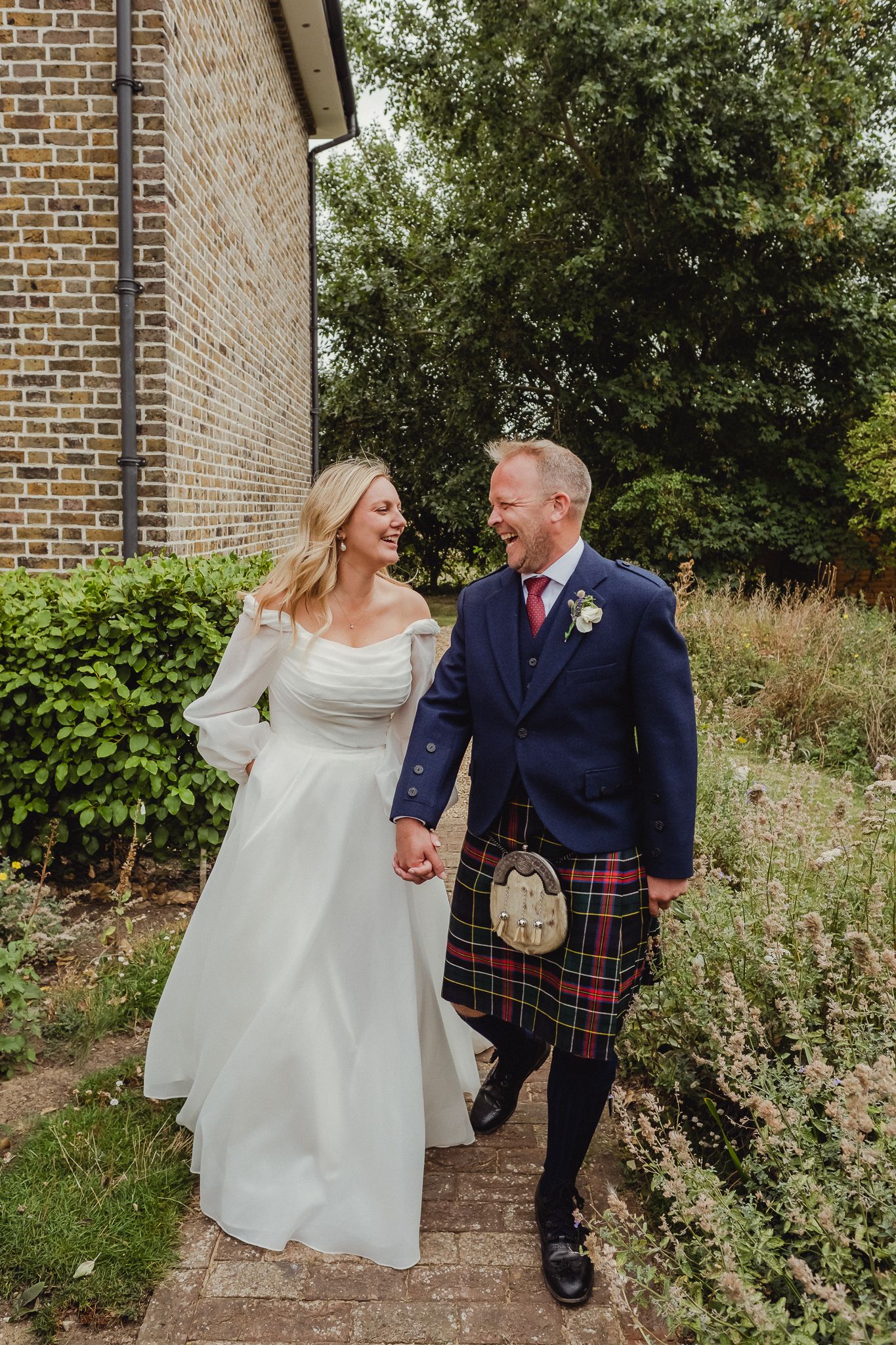 Candid wedding photos show couple walking and laughing together against brick wall and garden backdrop in Scottish attire.