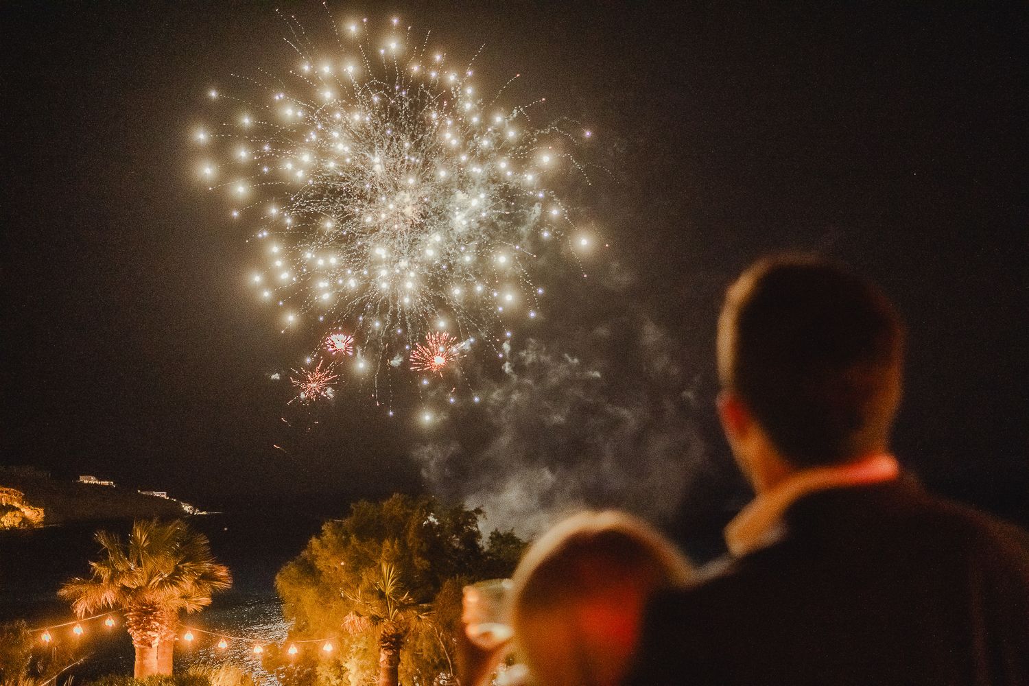 Silhouettes of spectators watch colorful fireworks exploding in the night sky over trees during an outdoor celebration.