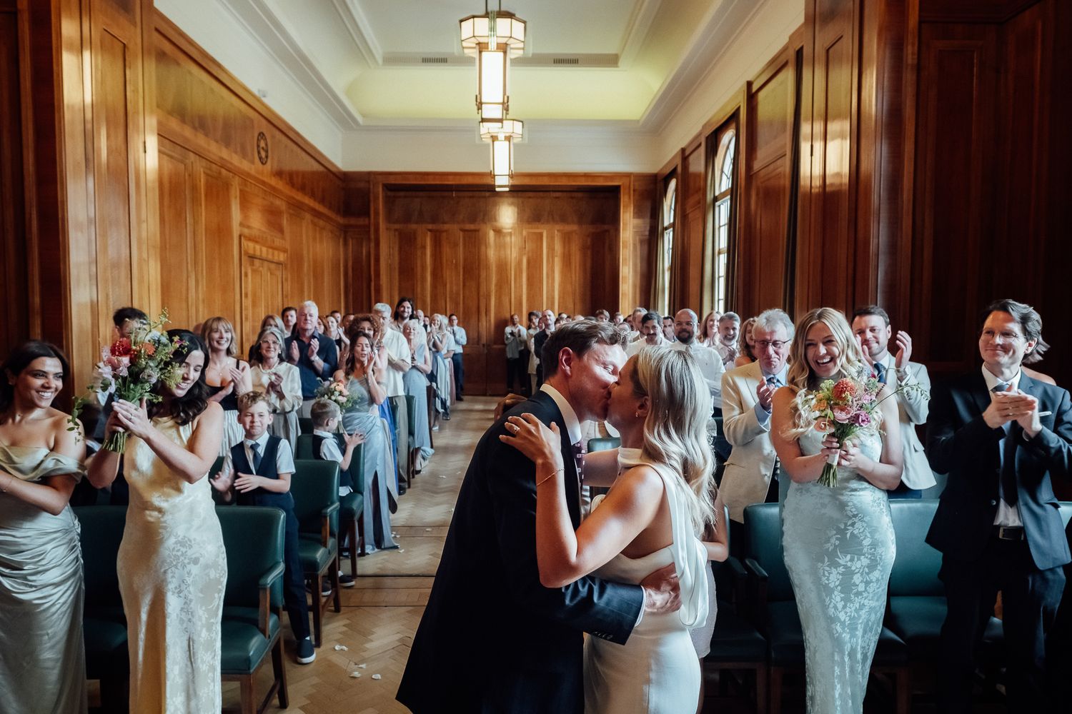 Newlywed couple shares a kiss in the aisle of a wood-paneled ceremony hall as smiling wedding guests in formal attire look on.