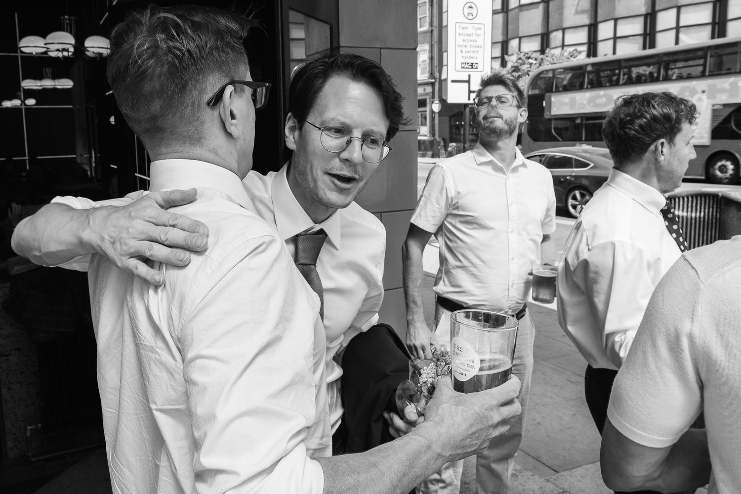 Black and white street photo of men in dress shirts chatting and holding beer glasses outside a pub, with a double-decker bus behind.