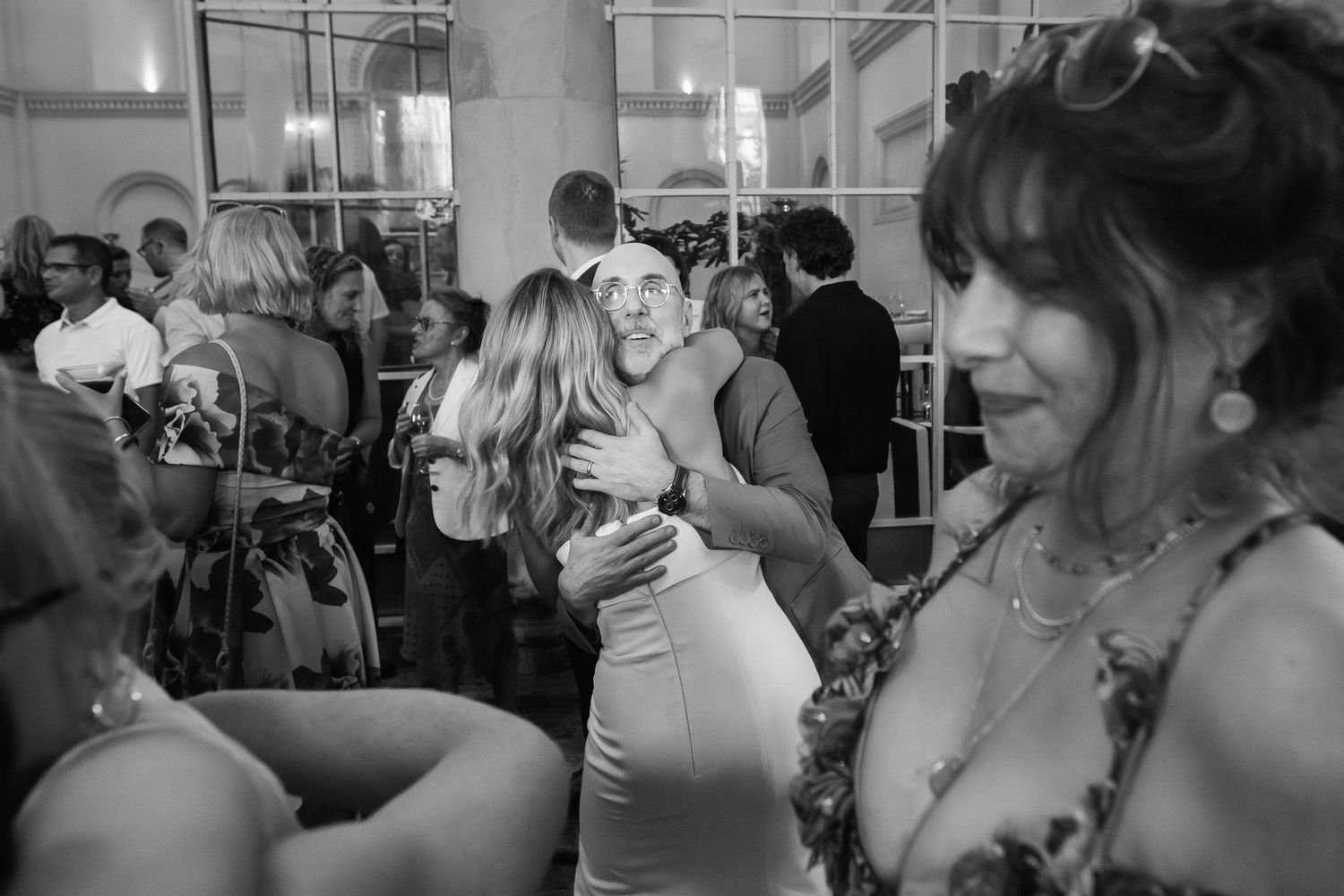 Black and white photo of wedding guests dancing and embracing on a crowded dance floor while a woman in a floral dress smiles in the foregro