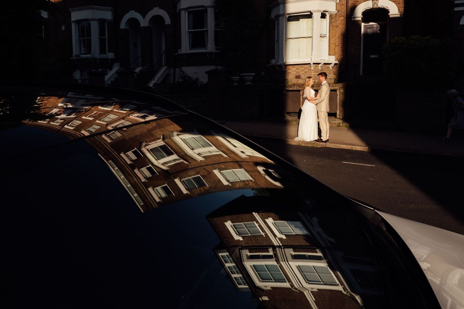 Bride and groom embrace on a sunlit city street while brick townhouses are reflected in the glossy dark roof of a parked car.