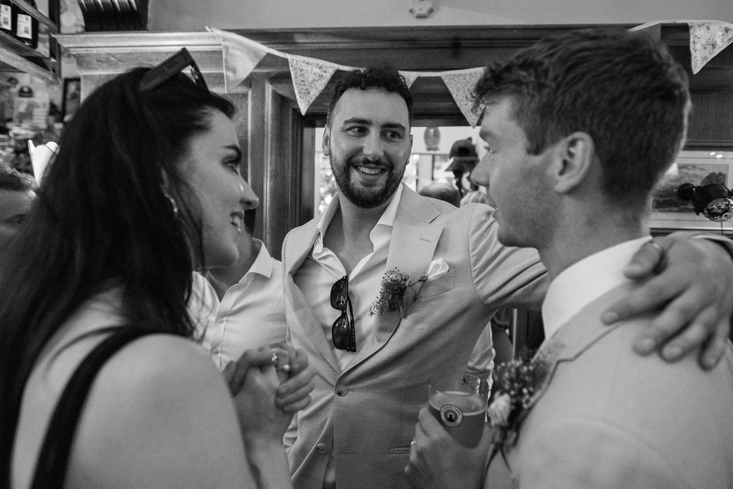 Three smiling wedding guests in formal attire embrace and laugh together under festive bunting in a black and white candid photo.