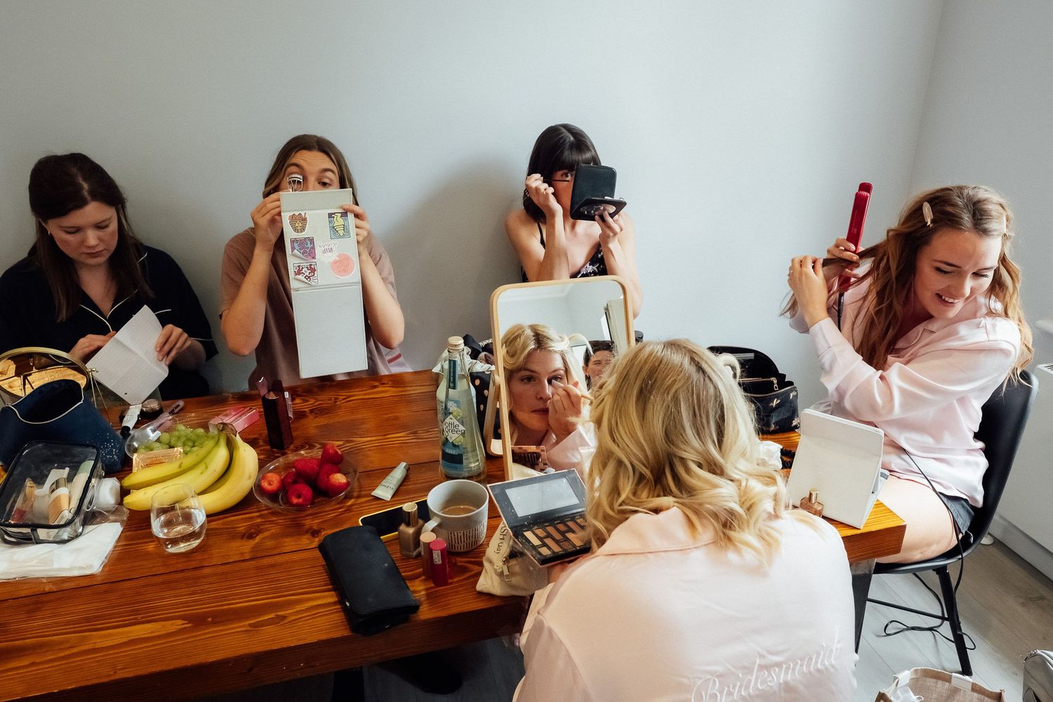 Five women getting ready around a wooden table with fruit, makeup, and a mirror, while one holds a curling iron and another reads a card.