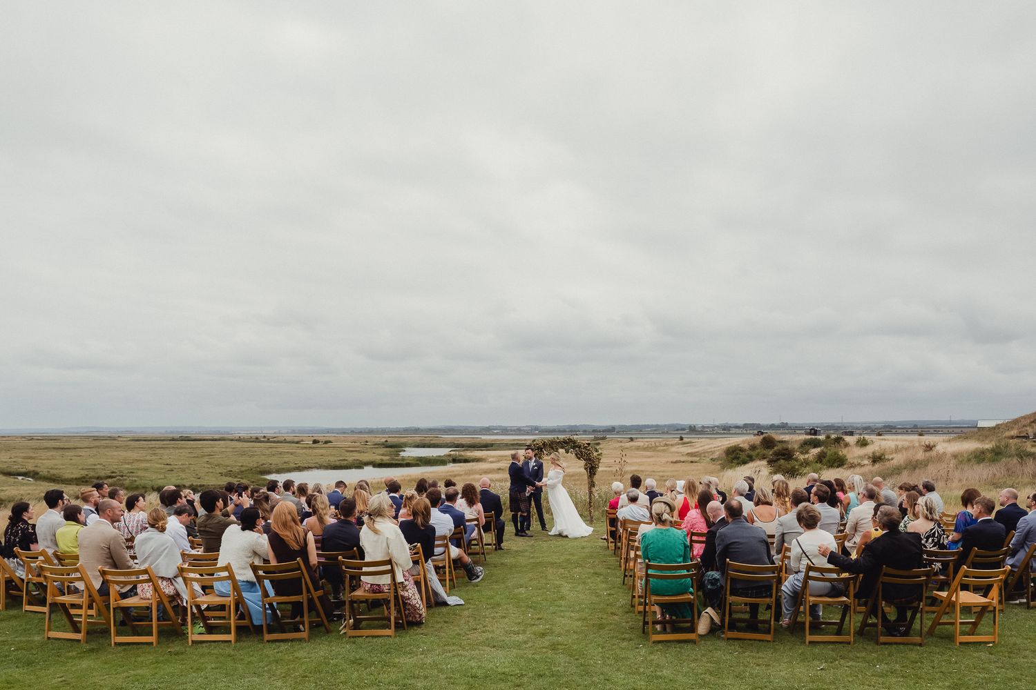 Outdoor wedding ceremony overlooking a scenic coastal landscape with guests seated on wooden chairs.