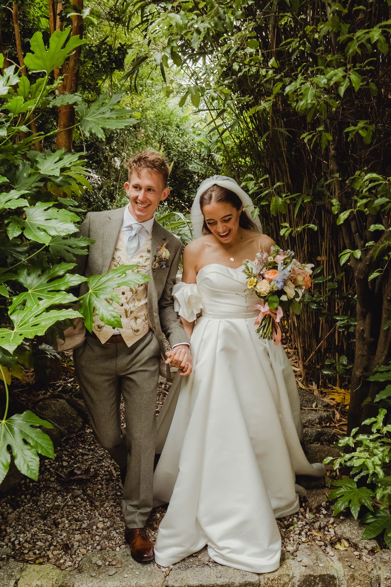 Couple shares joyful laughter in lush garden setting during outdoor wedding photoshoot.