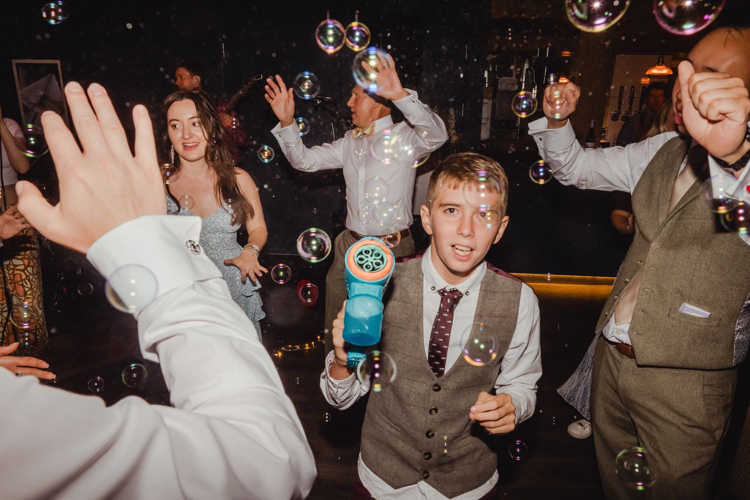 People dancing energetically at a dimly lit wedding reception with hands raised in celebration.