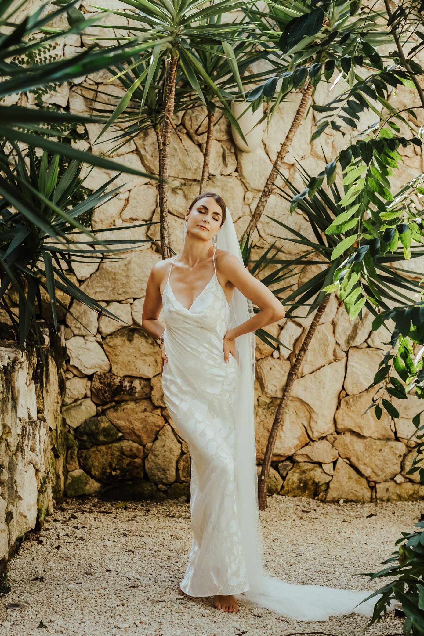 A figure in an elegant white wedding dress poses gracefully against a rustic stone wall surrounded by tropical foliage.