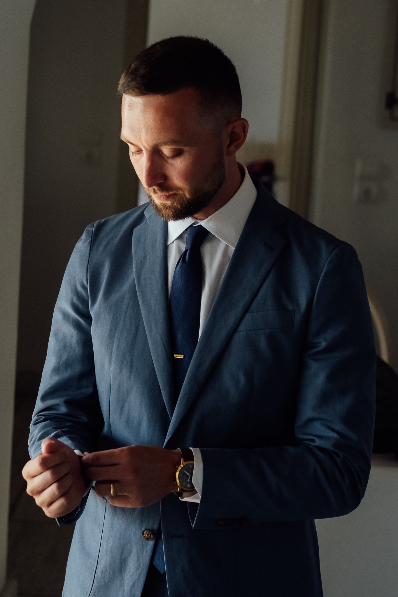 A well-dressed man in a navy blue suit adjusts his cufflinks while wearing a gold watch and navy tie.