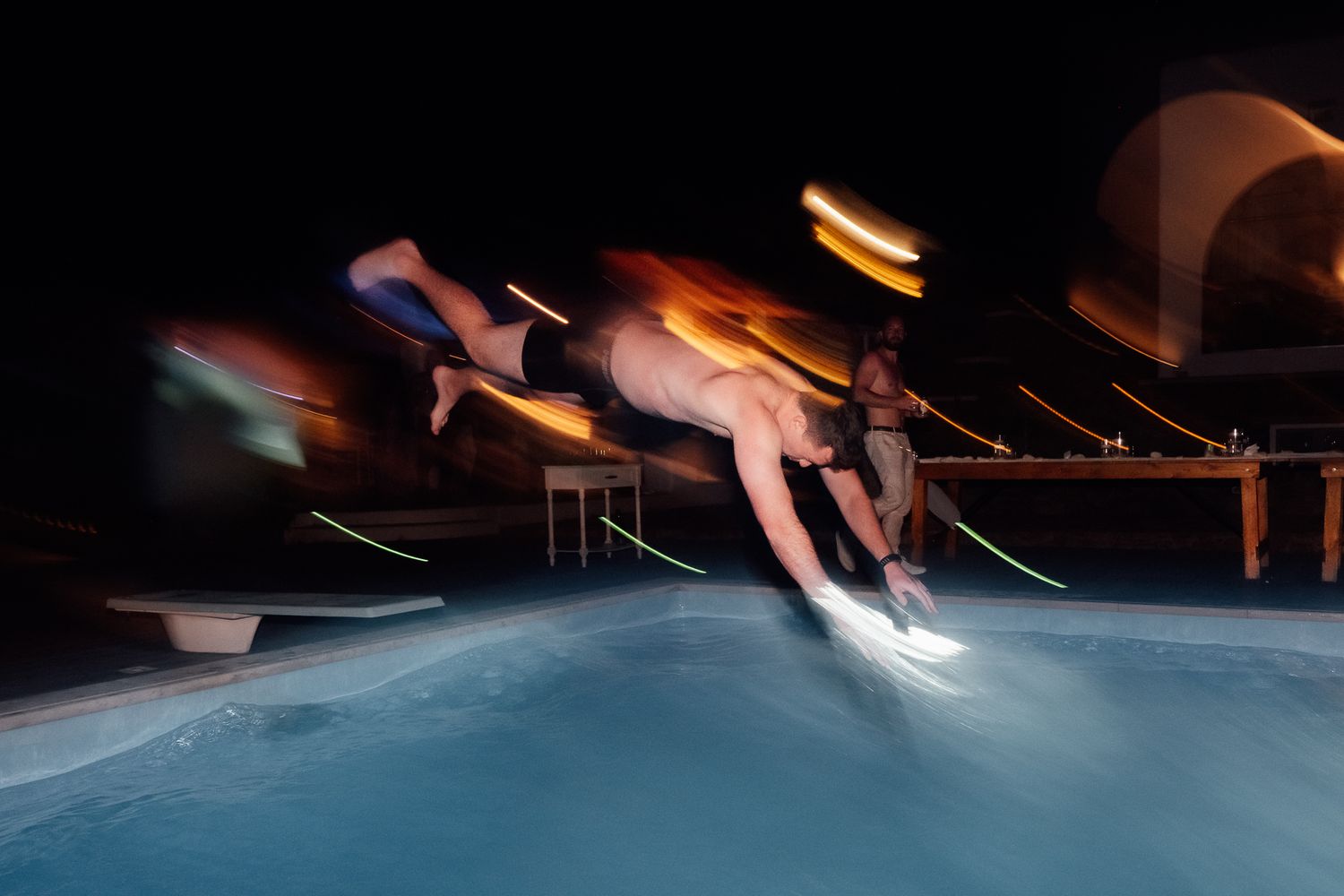 Man diving into illuminated pool at night with motion blur light trails captured in long exposure photography.