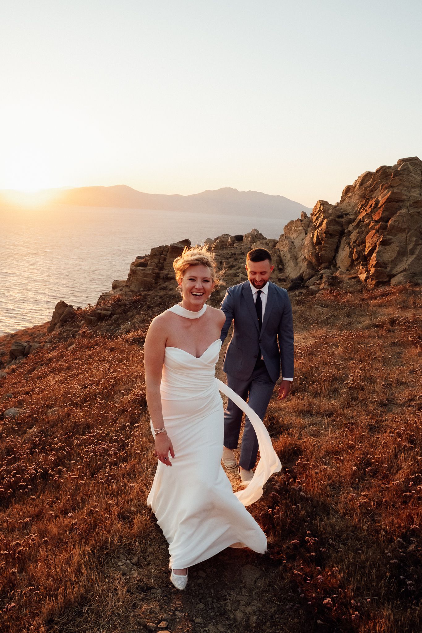 Bride and groom walk along rocky coastal cliffside at golden sunset, bride's white gown flowing in the breeze.