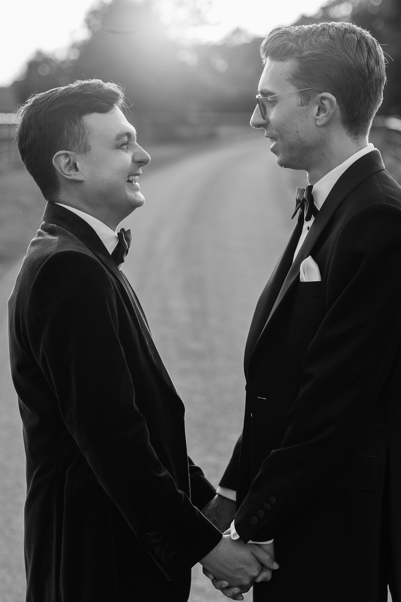 Black and white photo of two grooms in tuxedos holding hands and smiling at each other on their wedding day.