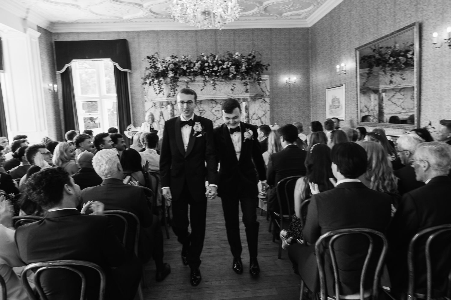 Two grooms walk down the aisle in tuxedos at an elegant black and white wedding ceremony.
