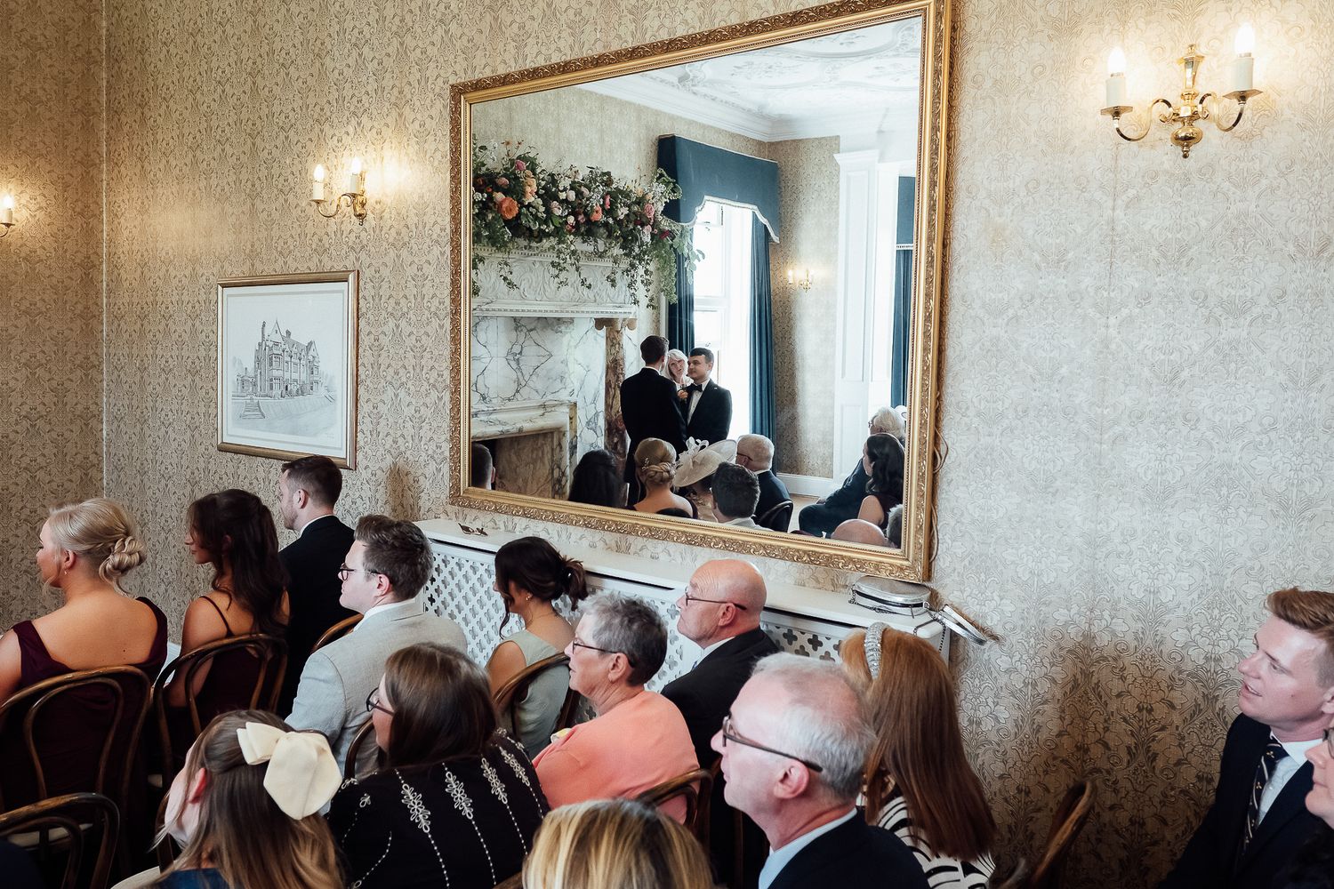 Wedding ceremony reflected in ornate gold mirror, guests seated in elegant wallpapered room with floral decorations.