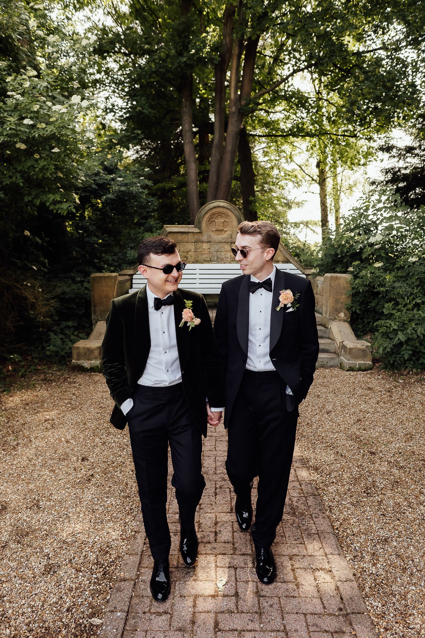 Two grooms in matching black tuxedos and sunglasses hold hands while walking through a garden path on their wedding day.