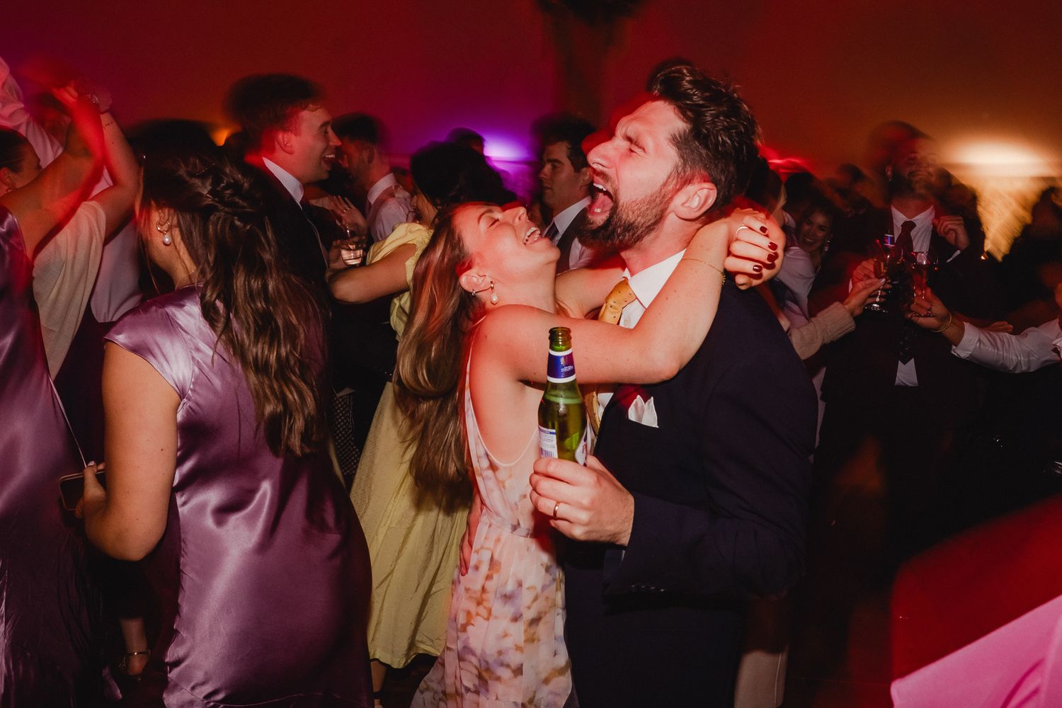 People dancing and celebrating at a lively party with colorful lighting and drinks in hand.