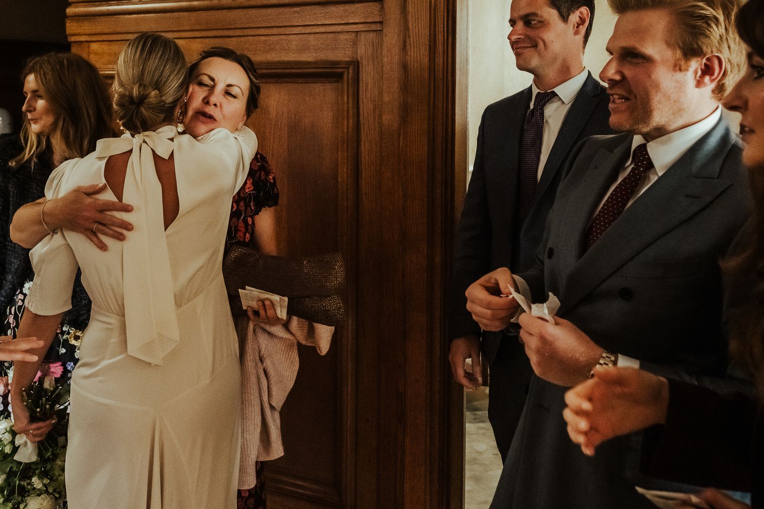 Wedding guests in formal attire viewing their phones as they congregate in a wood-paneled room at a reception.