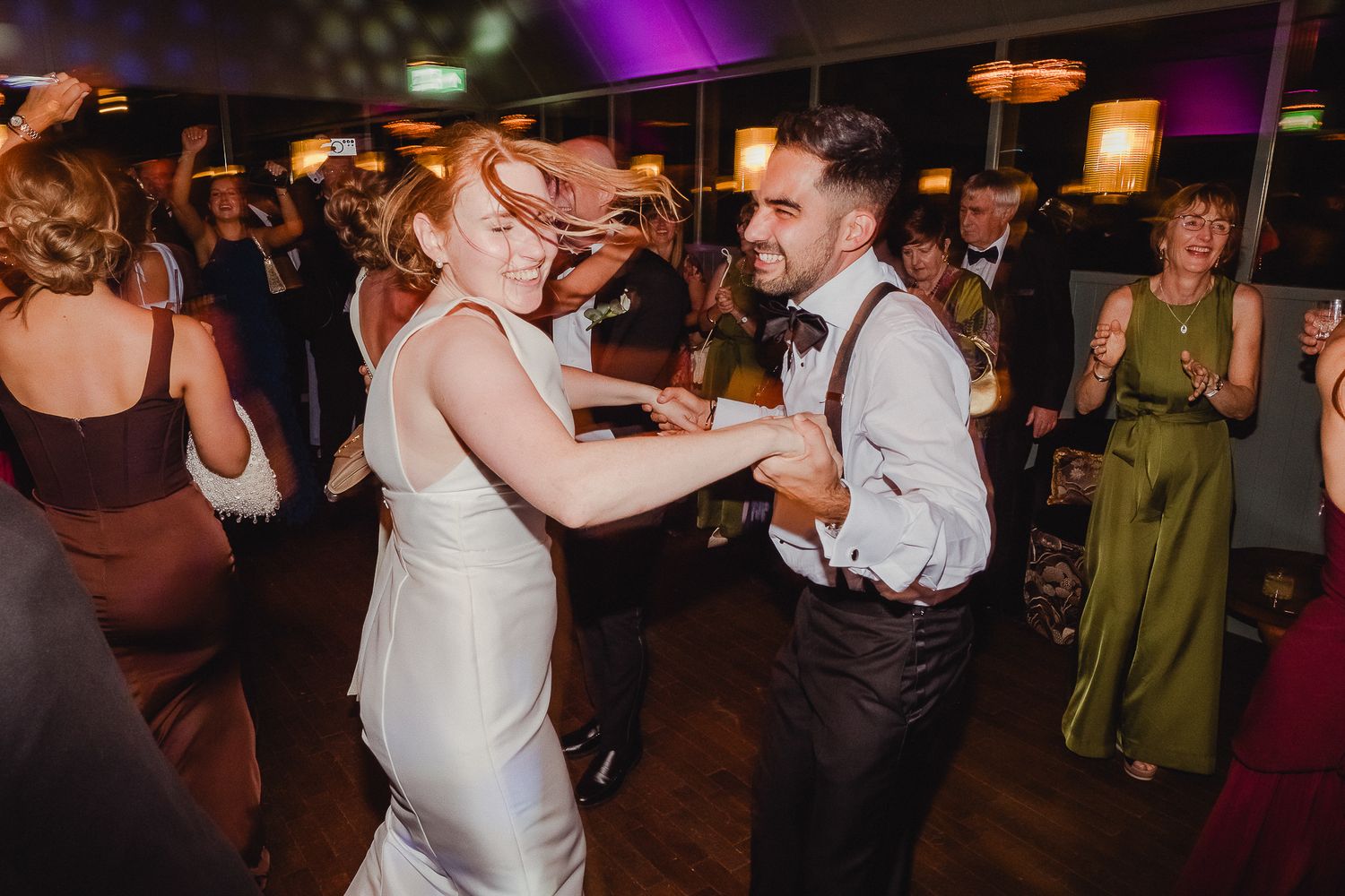 Wedding guests dance energetically on a dark dance floor illuminated by purple lighting at an evening reception.