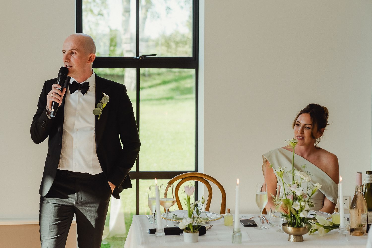 Wedding reception guests seated at tables while someone gives a speech at an elegant indoor venue.
