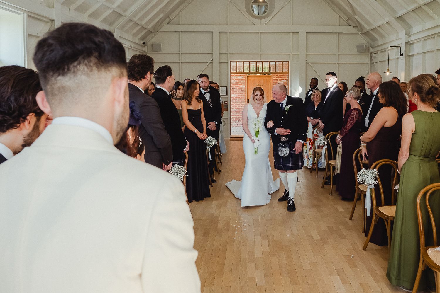 Wedding guests stand in a bright white reception hall with large windows and hardwood floors during a ceremony.