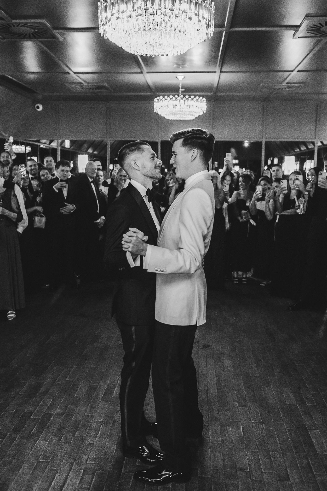 Couples dance together on a ballroom floor under crystal chandeliers during a formal evening event.