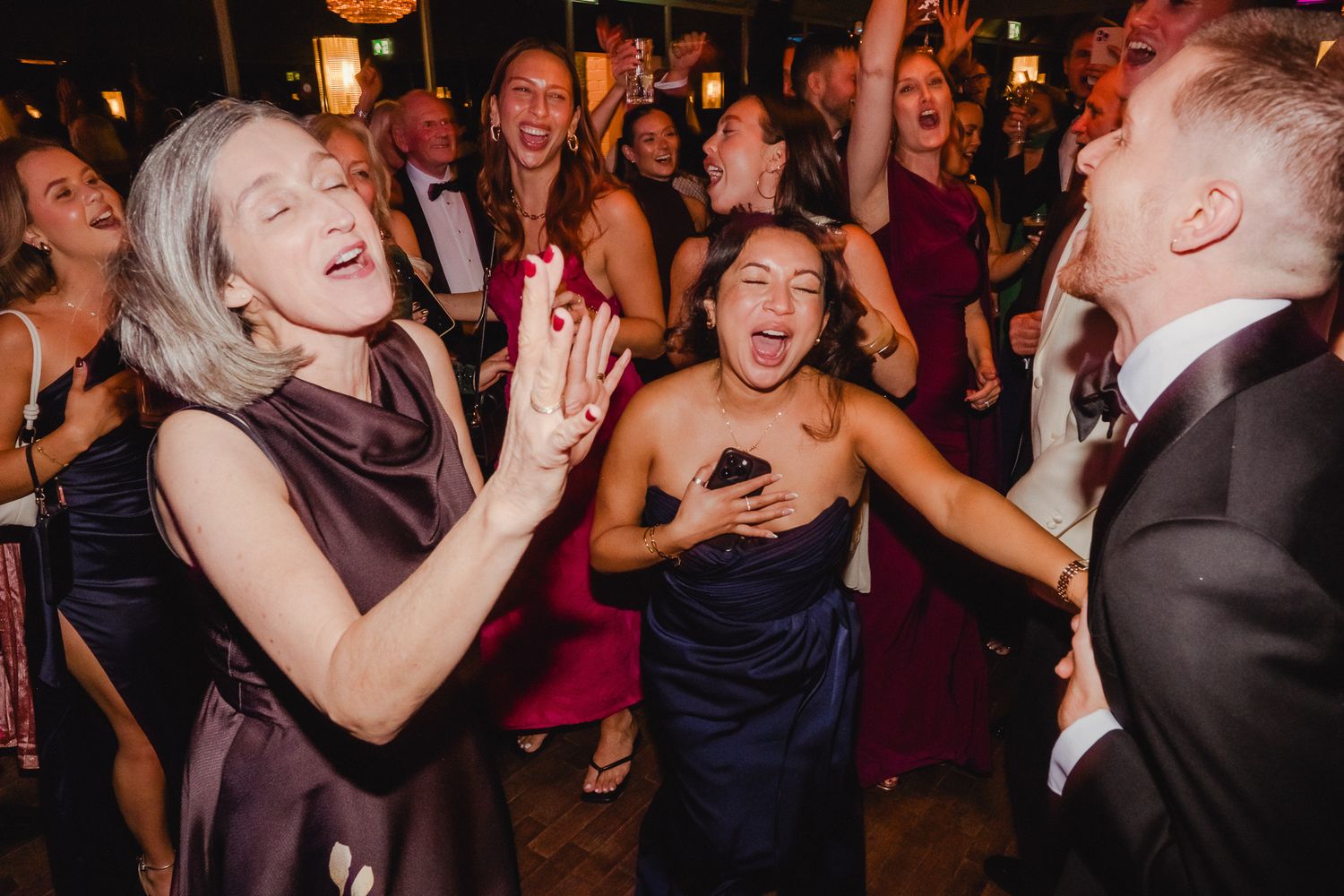 Wedding guests dancing and laughing together on a crowded dance floor at an evening reception celebration.