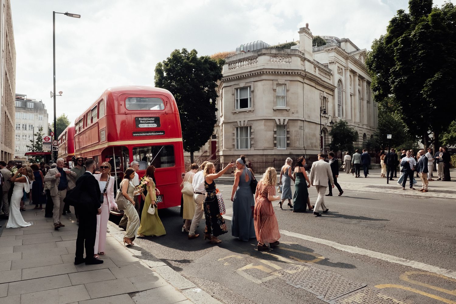 Group of people in colorful dresses and suits gather on a sunny city street beside a red double-decker bus and historic stone buildings.